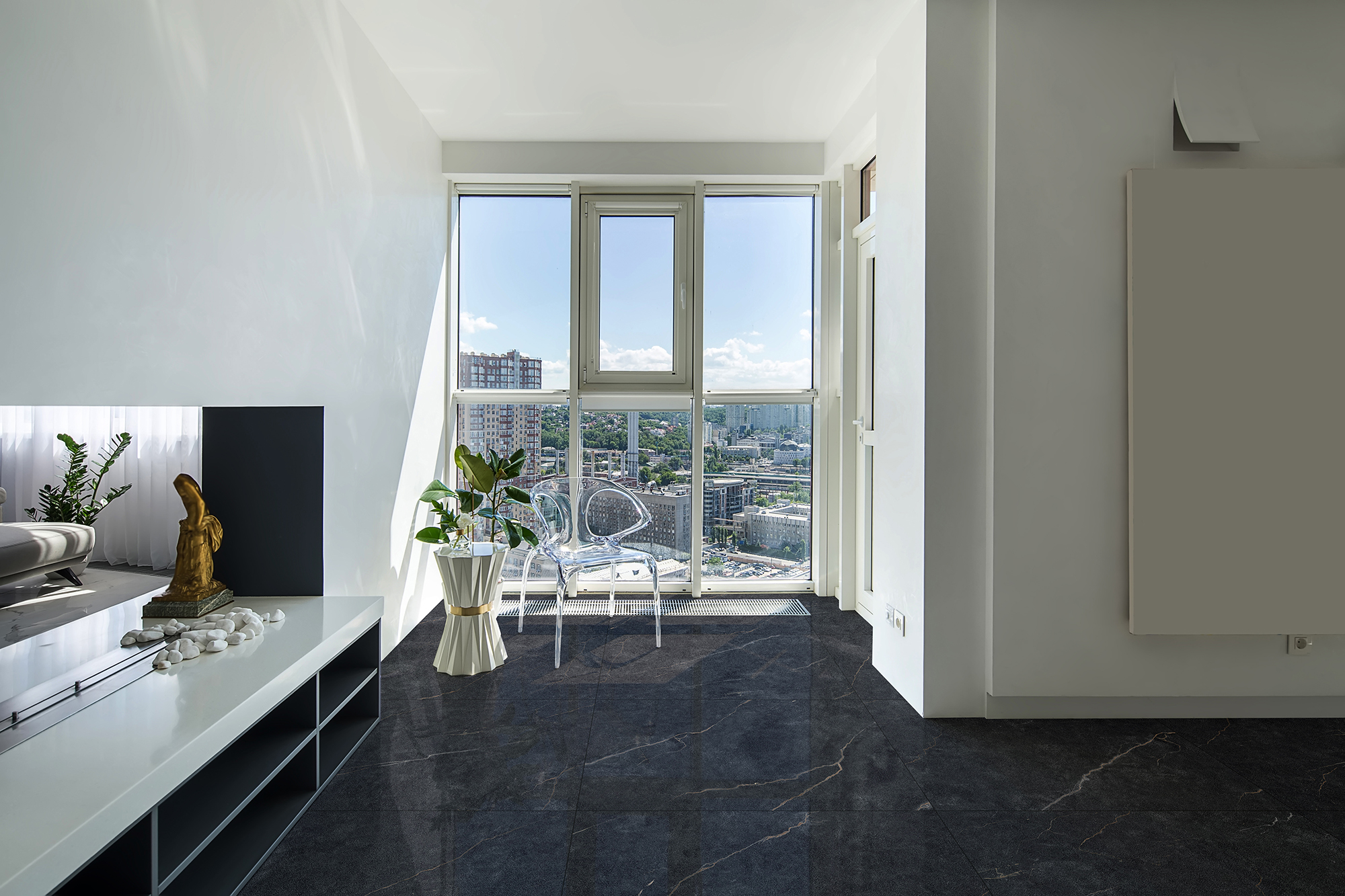 Interior in a modern style with white walls and light tiled floor. There is a stand with a white tabletop with a fireplace and stones and dark shelves, design stand flowers, glass chair, green plants.